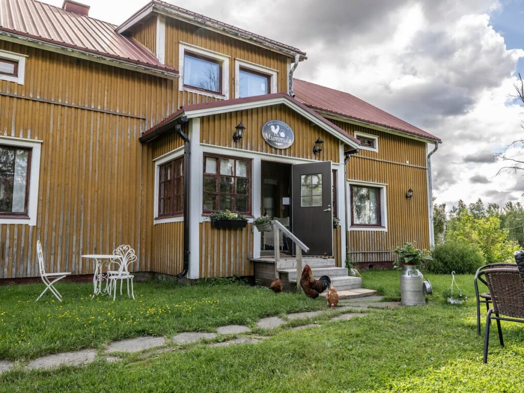 Kuva Maalaiskahvila Mummulasta. Kuvassa vanha keltainen puurakennus, jonka pihalla puutarhakalusteita ja kanoja.